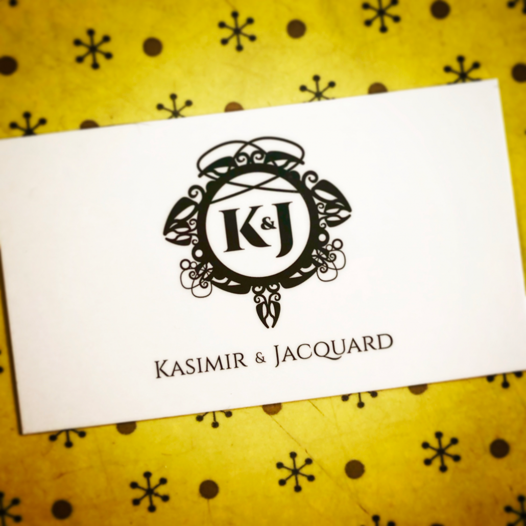 Carte d’affaire de Kasimir et Jacquard avec le logo noir et blanc en plein centre d’une carte blanche sur une table de travail jaune.
